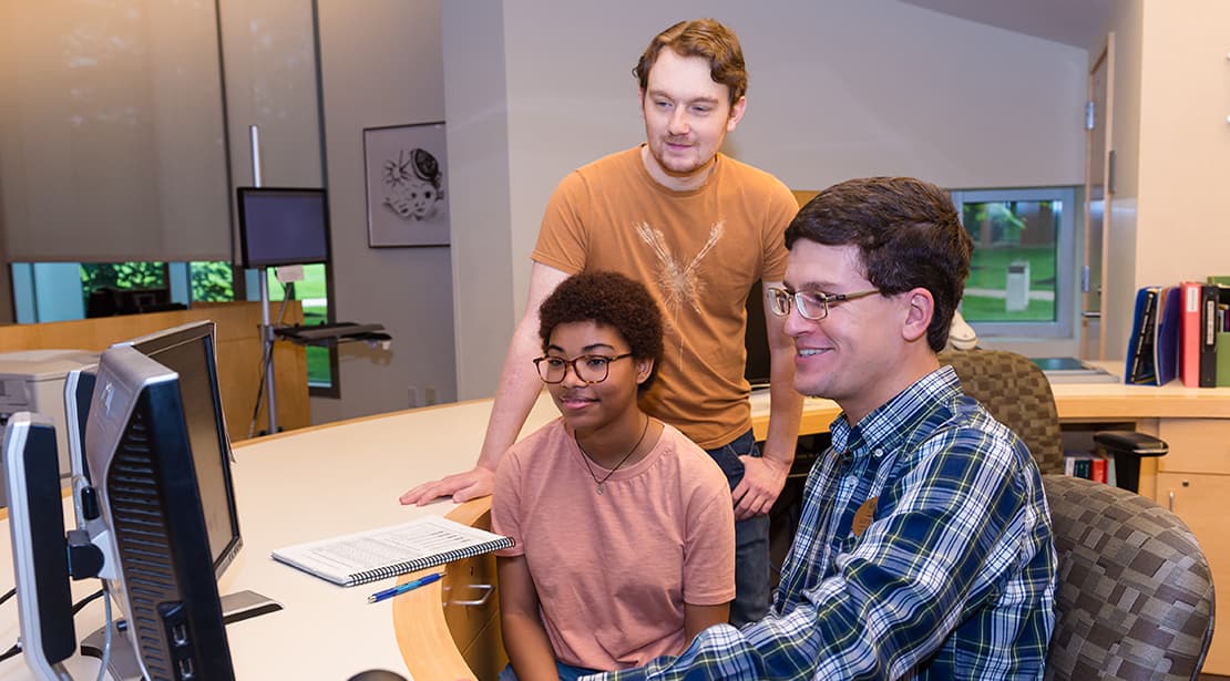 Three students sitting at desks smiling while looking at a laptop.