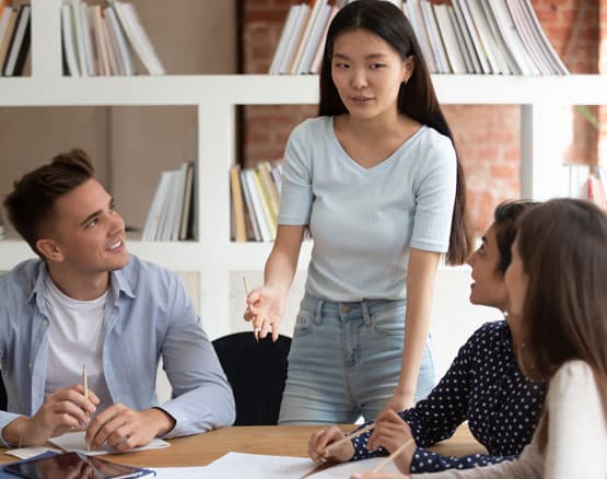Group of students listening as one young woman is speaking in classroom around a table with papers.