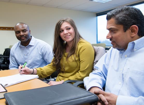 Three students sitting in a classroom; one is speaking, the other two are listening closely