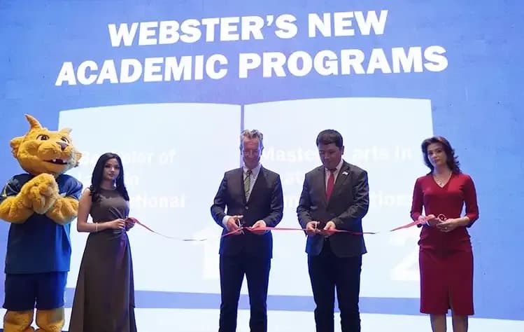 A ribbon-cutting ceremony featuring the Gorlok and University leadership on stage. They are launching two new academic programs, as indicated by the large blue backdrop reading "WEBSTER'S NEW ACADEMIC PROGRAMS."