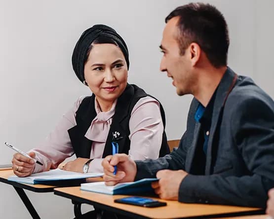 Female Webster student with braided hair talking during a class discussion with a fellow student behind her.