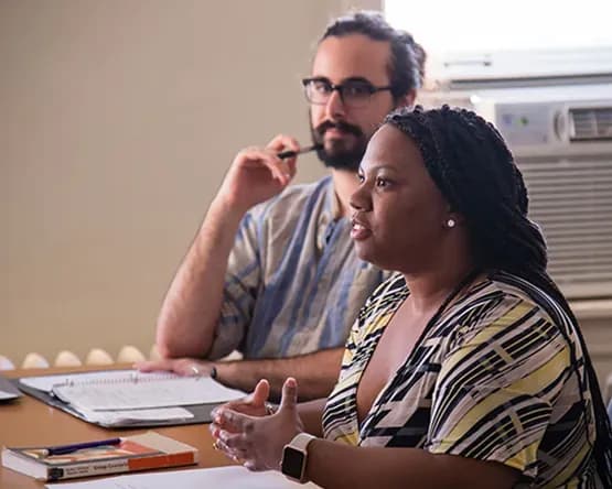 Two students sitting in a classroom; one is speaking, the other is listening closely